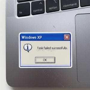 May include: A sticker featuring a Windows XP error message. The sticker has a blue border and displays the text "Windows XP" at the top. The message reads "Task failed successfully." with an "OK" button.