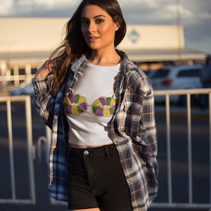 May include: A white crop top with a colorful floral design, paired with black shorts and a plaid shirt. The outfit is completed with a denim shirt. The overall style is casual and trendy.