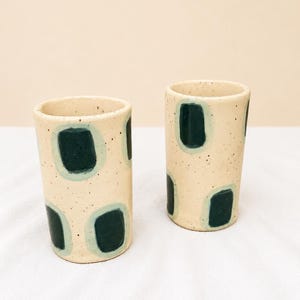 May include: Two cylindrical ceramic tumblers with a speckled cream-coloured glaze. Each vessel features a repeating pattern of dark green rectangular shapes with rounded corners, outlined by a soft light blue border around each dark green mark.