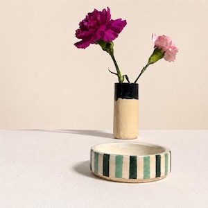 Peut inclure: Un petit vase cylindrique avec un dessus noir et un corps beige contient deux fleurs : un œillet magenta foncé et un œillet rose clair. Un bol rond en céramique avec des rayures vertes et turquoise est devant le vase.