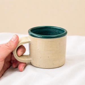 May include: A ceramic mug with a speckled cream-coloured exterior and a smooth, dark teal interior rim. The mug features a sturdy, thick handle and a cylindrical body, showcasing a handcrafted aesthetic with subtle texture and natural colour variations.