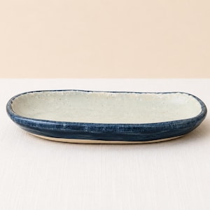 Peut inclure: Un plat ovale en céramique avec un intérieur blanc cassé moucheté et un extérieur émaillé bleu profond. Le bord présente une texture irrégulière façonnée à la main, et la base du plat montre une finition en argile naturelle non émaillée.