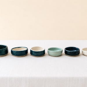 May include: A set of six small ceramic bowls arranged in a horizontal row. The bowls feature varying shades of blue, teal, and off-white, with smooth, rounded edges and a glossy finish on the interior and exterior surfaces of each individual vessel.