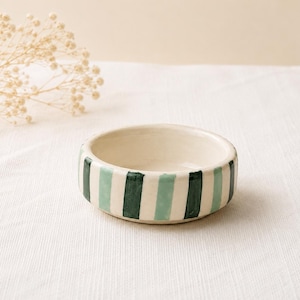 May include: A small, round ceramic bowl featuring a hand-painted pattern of alternating vertical stripes in dark forest green and light mint green. The interior of the bowl is a solid off-white colour, and the exterior base is also finished in off-white.