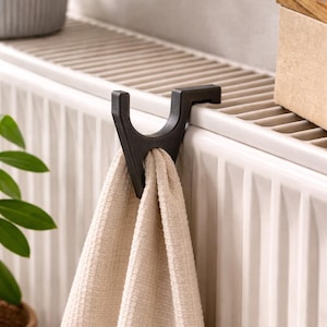 Può includere: Un gancio portasciugamani marrone scuro è attaccato a un radiatore bianco, che tiene un asciugamano beige con strisce grigie. Il gancio ha un design curvo unico. Una pianta in vaso è visibile sullo sfondo.