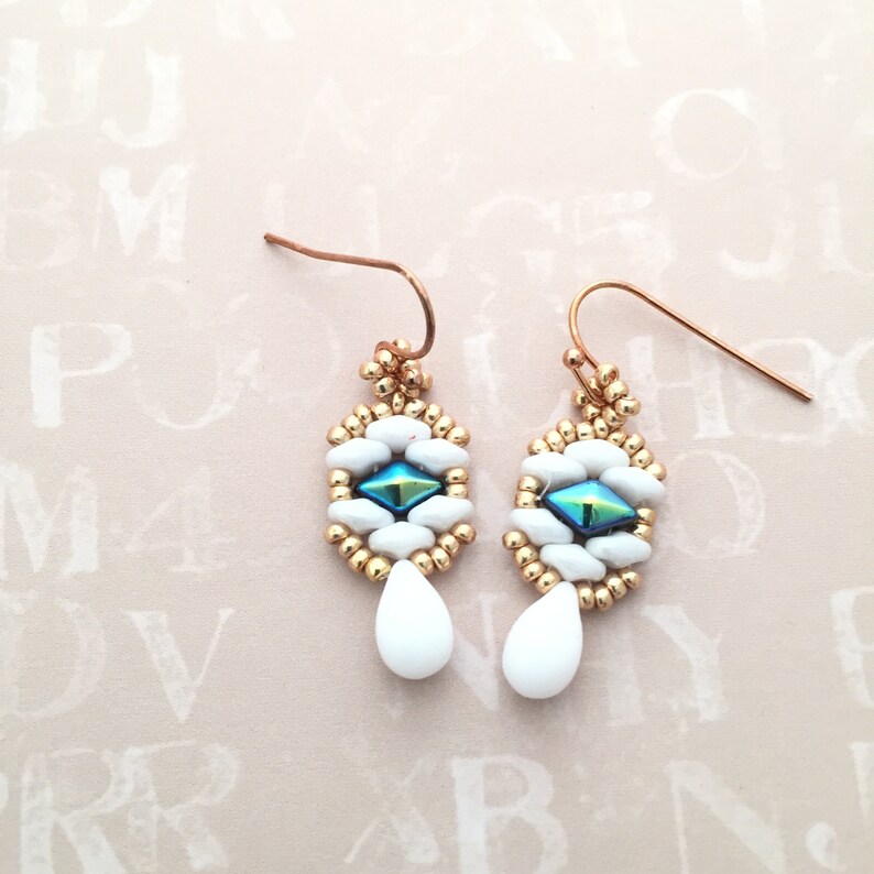 May include: A pair of gold earrings with white and blue beads. The earrings have a teardrop shape with a small blue diamond-shaped bead in the center.
