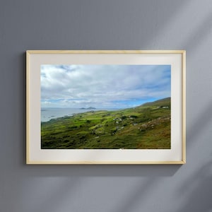May include: A framed landscape photograph of a coastal scene. The image features rolling green hills, a body of water, and a cloudy sky. Small buildings are scattered across the landscape. The frame is a light wood color.