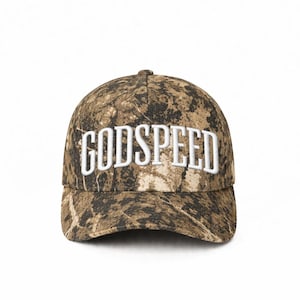Op de afbeelding: Een camouflage baseballpet met het woord "GODSPEED" in het wit geborduurd. De pet heeft een bruin en beige patroon en een gebogen klep.