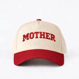 Pode incluir: Boné de beisebol com coroa creme e aba vermelha. A palavra "MOTHER" é bordada em vermelho na frente do boné. O boné tem uma aba curva e um botão na parte superior.