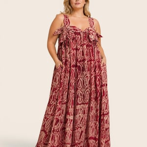 May include: A floor-length red dress with a paisley pattern and pockets. The dress features wide shoulder straps with bow details. The fabric has a textured appearance. The model is wearing the dress.
