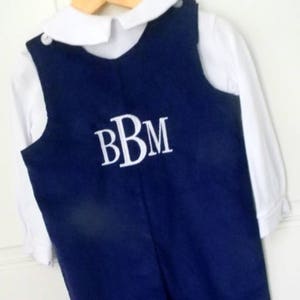 May include: A navy blue overall with white straps and a white button closure. The overall has a white embroidered monogram "BBM" on the front.