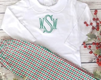 May include: A white long-sleeved shirt with a Peter Pan collar and green embroidered initials, paired with red and green plaid pants. The outfit is displayed on a light wood surface, with decorative holly and berries.