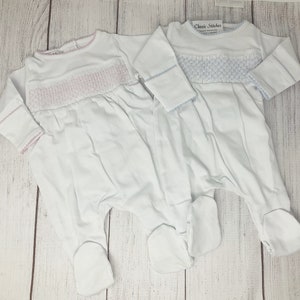May include: Two white baby footed pajamas with long sleeves. One has pink smocking detail, the other has blue smocking. Both have matching trim around the neckline and cuffs. The pajamas are displayed on a light wood grain surface.