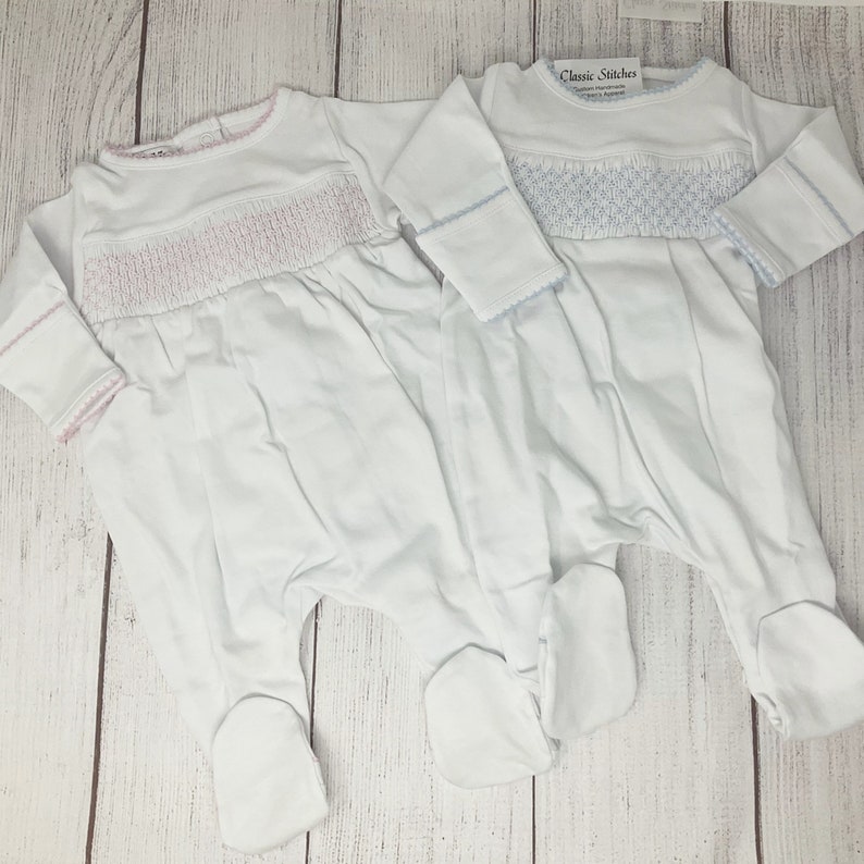 May include: Two white baby footed pajamas with long sleeves. One has pink smocking detail, the other has blue smocking. Both have matching trim around the neckline and cuffs. The pajamas are displayed on a light wood grain surface.