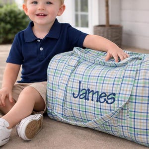 May include: A blue, green, and tan plaid duffel bag with the name "James" embroidered in blue. The bag has two handles and a zipper closure. The bag is sitting on a concrete surface.