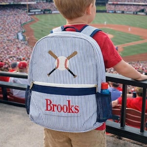 May include: A blue and white striped backpack with a baseball theme. The backpack features an embroidered baseball and crossed bats, a navy blue pocket, and the name "Brooks" in red. A water bottle is in the side pocket.