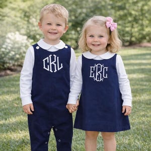 May include: Two children wearing matching navy blue outfits with white collars and long-sleeved shirts. The boy wears a romper with the initials "CKL", and the girl wears a dress with the initials "E&C". Both outfits are embroidered with white lettering.