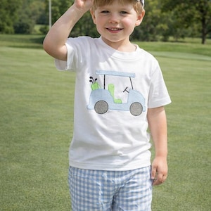 May include: A young child wearing a white t-shirt with a golf cart design in light blue, green, and gray. The child is also wearing blue and white checkered shorts and a white baseball cap. The background is a green golf course.