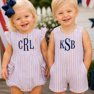 May include: Two children wearing matching red, white, and blue striped rompers. The girl's romper has ruffled sleeves and pockets, and a navy bow. Both outfits are monogrammed with navy letters. The children are smiling.