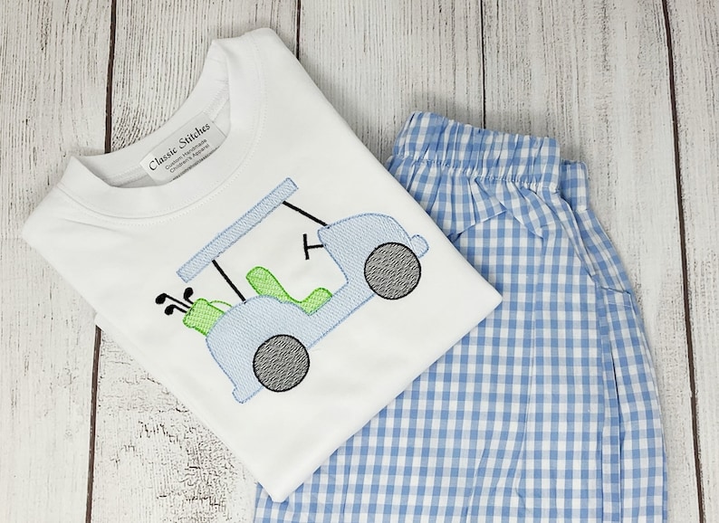May include: White t-shirt with a light blue golf cart applique, green seats, and black wheels. The shirt is paired with blue and white gingham shorts. The shirt has the text "Classic Stitches" on the label.
