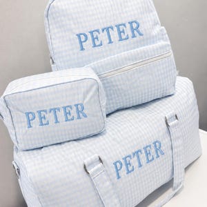 May include: A set of light blue and white gingham travel bags, including a backpack, a duffel bag, and a cosmetic bag. Each bag has the name "PETER" embroidered in light blue. The bags are stacked on top of each other.