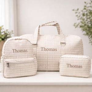 May include: A set of three beige and white gingham travel bags, including a backpack, a duffel bag, and a cosmetic bag. Each bag is embroidered with the name "Thomas" in a matching beige thread. The bags are arranged on a white surface.