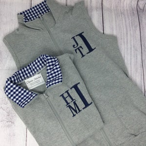 May include: Two gray fleece vests with blue and white gingham collars. The vests have a zipper closure and are personalized with embroidered initials. The vest on the left has the initials "HM" and the vest on the right has the initials "JT".