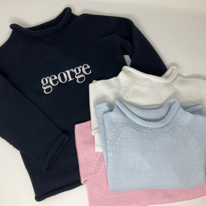 May include: A stack of knit sweaters in navy, white, light blue, and pink. The navy sweater has the name "george" embroidered in white. The sweaters have a mock neck and long sleeves, suitable for children's wear.