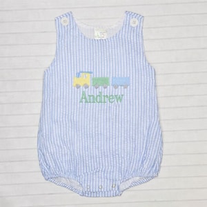 May include: A blue and white striped seersucker romper with a train applique in yellow, green, and blue. The name "Andrew" is embroidered in green below the train. The romper has button closures at the shoulders and bottom.