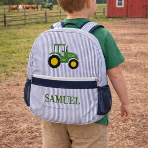May include: A blue and white striped backpack with a green tractor design and the name "SAMUEL" embroidered in green. The backpack has navy blue accents and mesh side pockets. The child is wearing a green shirt and khaki shorts.