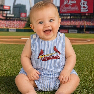 May include: A smiling baby wearing a blue and white striped romper with the St. Louis Cardinals logo, sitting on a baseball field. The background includes the stadium and the Gateway Arch.