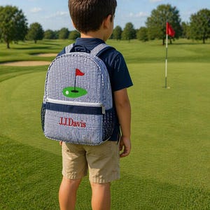 May include: A blue and white striped backpack with a golf-themed design. The backpack features a green golf course with a red flag and a golf ball. The name "JJ Davis" is embroidered in red. The backpack is worn by a child on a golf course.