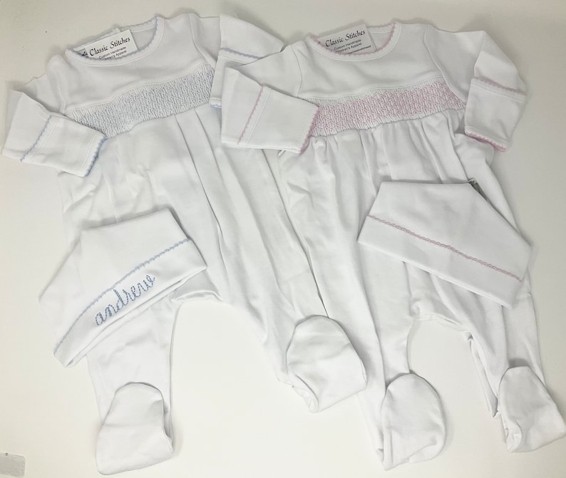May include: Two white baby footed sleepers with matching hats. One sleeper has blue trim and smocking, the other has pink trim and smocking. The hat of the blue-trimmed sleeper is embroidered with the name "Andrew". The sleepers have long sleeves.