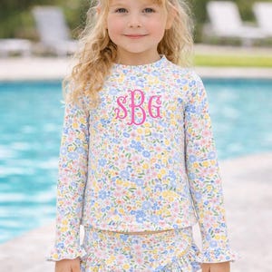 May include: A young girl wearing a floral print swimsuit with long sleeves and ruffled edges. The swimsuit has a light blue, yellow, and pink floral pattern. The initials "SBG" are embroidered in pink on the chest. She is wearing a large pink bow in her hair.