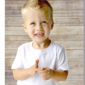 May include: A smiling child with blue eyes and a blonde mohawk hairstyle is wearing a white t-shirt. The child is holding up their fingers. The background is a light brown wooden wall.