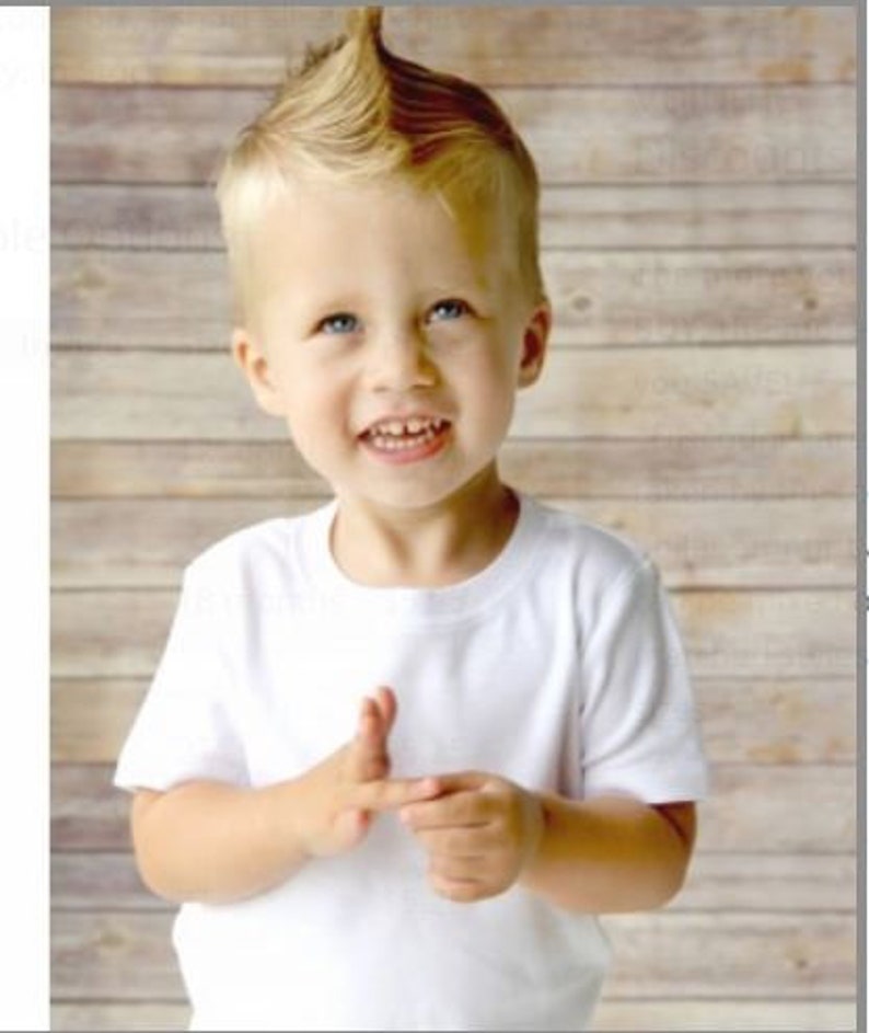 May include: A smiling child with blue eyes and a blonde mohawk hairstyle is wearing a white t-shirt. The child is holding up their fingers. The background is a light brown wooden wall.