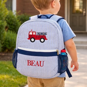 May include: A blue and white striped backpack with a red fire truck design and the name "BEAU" embroidered in red. The backpack has a navy blue trim, a side mesh pocket, and a water bottle. The child is wearing a blue shirt and khaki shorts.