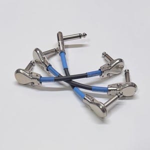 Puede incluir: Tres cables de conexión cortos con secciones de goma negras y azules y conectores metálicos plateados en ángulo recto. Cada cable cuenta con un extremo de enchufe metálico y una funda azul cerca del conector, con texto impreso en las partes azules.