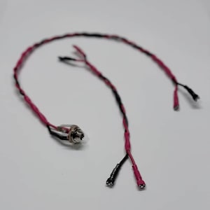 May include: A close-up of a black and pink wire assembly with a silver jack. The wires are twisted together and have a heat shrink tubing. The assembly is on a white surface.