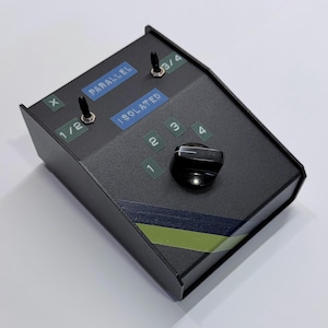 May include: A black electronic device with multiple switches and a rotary knob. The device has labels reading "PARALLEL" and "ISOLATED". Additional labels indicate "1/2" and "3/4". A green and blue stripe design is visible on the side.
