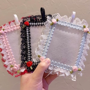 May include: Three decorative frames with lace trim and small rose accents. The frames are in pink, black and white gingham, and light blue. Each frame has a ribbon loop for hanging.