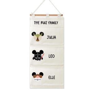 Puede incluir: Un organizador colgante color crema con tres bolsillos, cada uno con un diseño de personaje de Star Wars. El bolsillo superior dice "THE RUIZ FAMILY". Debajo están los nombres "JULIA", "LEO" y "ELLE". El organizador está suspendido por una barra de madera y un cordón blanco.