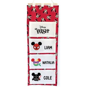 May include: Red fabric wall organizer with four pockets. The top is patterned with Minnie Mouse faces. The pockets feature Disney characters and names: "WISH", "LIAM", "NATALIA", and "COLE". The organizer hangs from a wooden rod.