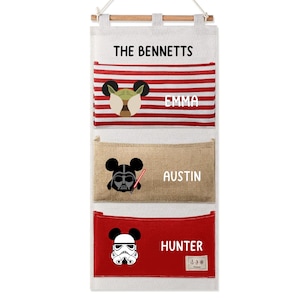 May include: A hanging wall organiser with three fabric pockets. The top pocket has red and white stripes with a Yoda-inspired icon and the name EMMA. The middle pocket is tan with a Darth Vader icon and the name AUSTIN. The bottom pocket is red with a Stormtrooper icon and the name HUNTER. The top header reads THE BENNETTS.