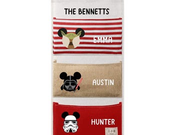 Puede incluir: Un organizador de pared colgante con tres bolsillos de tela. El bolsillo superior tiene rayas rojas y blancas con un icono inspirado en Yoda y el nombre EMMA. El bolsillo central es beige con un icono de Darth Vader y el nombre AUSTIN. El bolsillo inferior es rojo con un icono de Stormtrooper y el nombre HUNTER. El encabezado dice THE BENNETTS.