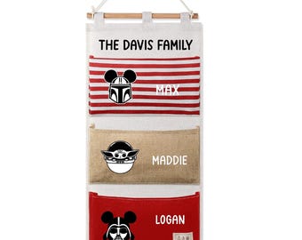 May include: A wall hanging organizer with three pockets. The top pocket has red and white stripes and a black and white Mandalorian helmet design. The middle pocket is tan with a Baby Yoda design. The bottom pocket is red with a Darth Vader design. The text "THE DAVIS FAMILY" is at the top.