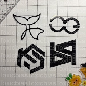 May include: Four black vinyl decals on a white grid background. The designs include a whale tail outline, a stylized infinity symbol, a geometric hexagon shape with internal lines, and a bold angular letter design. The grid features a ruler edge.