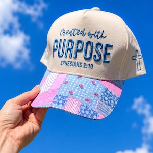K&ouml;nnte beinhalten: Eine beige Baseballkappe mit blauer Stickerei, die den Text Created with PURPOSE EPHESIANS 2:10 zeigt. Der Schirm ist im Patchwork-Stil mit blauen und rosafarbenen Blumen- und Streifenmustern gestaltet und an der Seite befindet sich ein Kreuz.