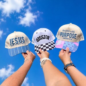K&ouml;nnte beinhalten: Drei Baseballkappen vor einem blauen Himmel. Die linke Kappe ist cremefarben mit blauem Text JESUS THE WAY THE TRUTH THE LIFE und blau gestreiftem Schirm. Die mittlere Kappe ist wei&szlig; mit schwarzem Text BLESSED und schwarz-wei&szlig; kariertem Schirm. Die rechte Kappe ist cremefarben mit blauem Text Created with PURPOSE EPHESIANS 2:10 und einem blau-rosa gebl&uuml;mten Patchwork-Schirm.