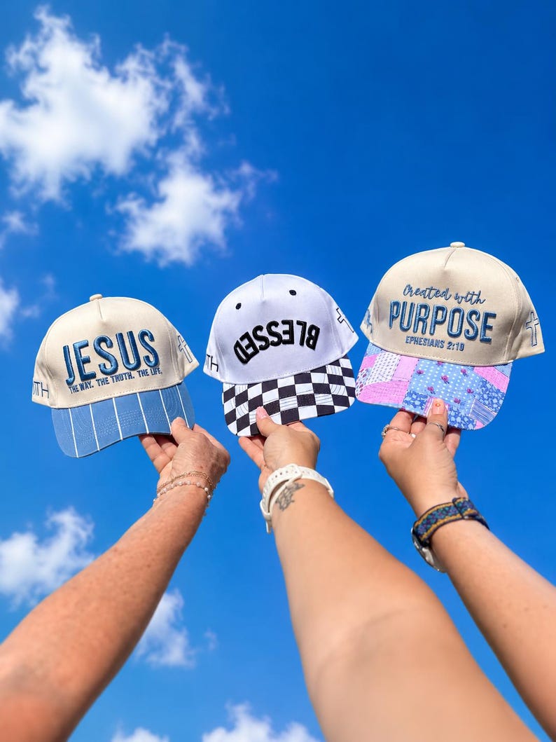 K&ouml;nnte beinhalten: Drei Baseballkappen vor einem blauen Himmel. Die linke Kappe ist cremefarben mit blauem Text JESUS THE WAY THE TRUTH THE LIFE und blau gestreiftem Schirm. Die mittlere Kappe ist wei&szlig; mit schwarzem Text BLESSED und schwarz-wei&szlig; kariertem Schirm. Die rechte Kappe ist cremefarben mit blauem Text Created with PURPOSE EPHESIANS 2:10 und einem blau-rosa gebl&uuml;mten Patchwork-Schirm.